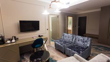 A hotel room with a flat-screen TV, a desk with a coffee maker, a blue chair, a patterned sofa, and a small table.