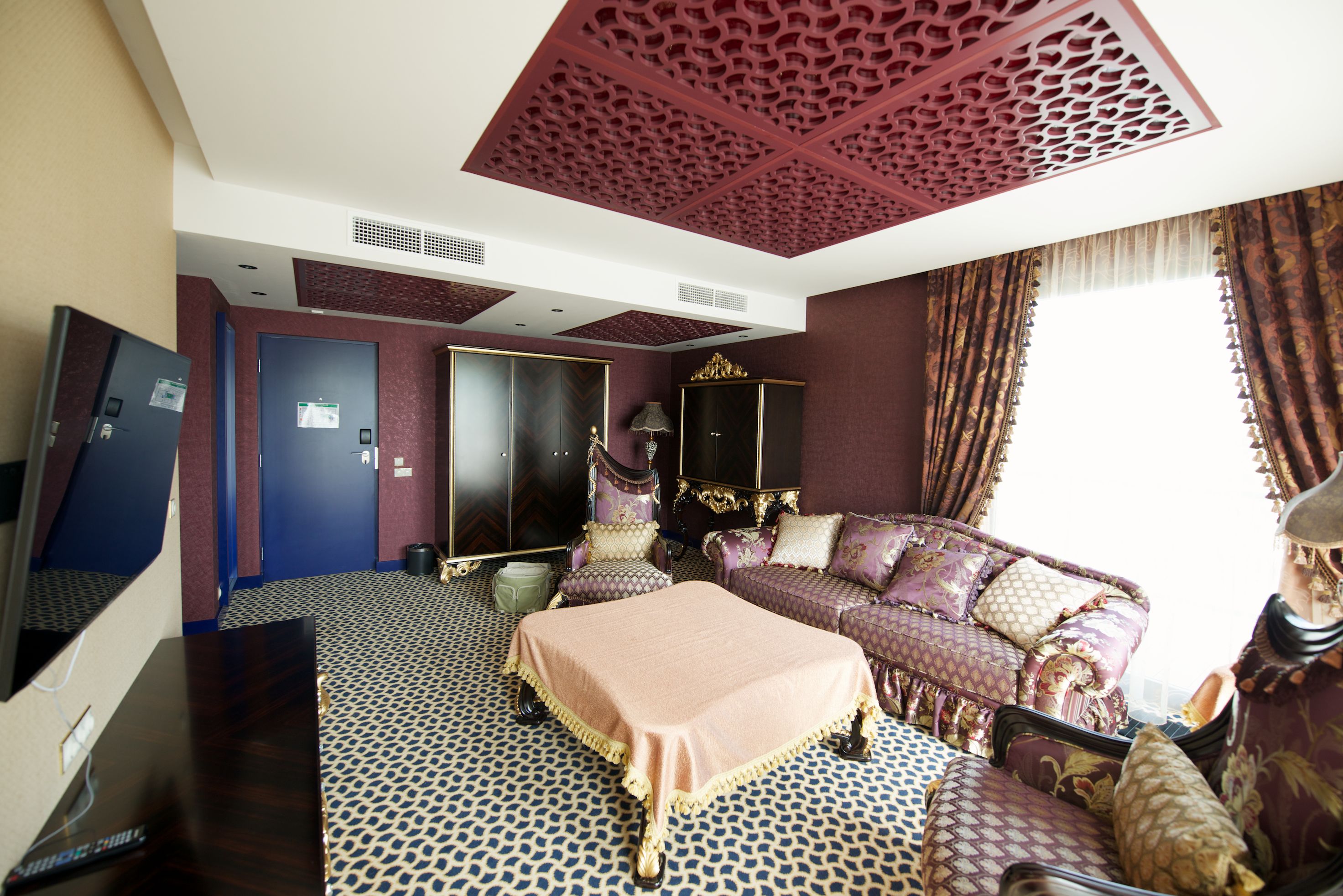 A hotel room with a patterned floor, a bed with a pink bedspread, a sofa with patterned cushions, and a decorative ceiling.