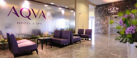 Un hall con mostrador de recepción, asientos morados, piso de mármol y una pared con el rótulo 'AQVA HOTEL & SPA'.