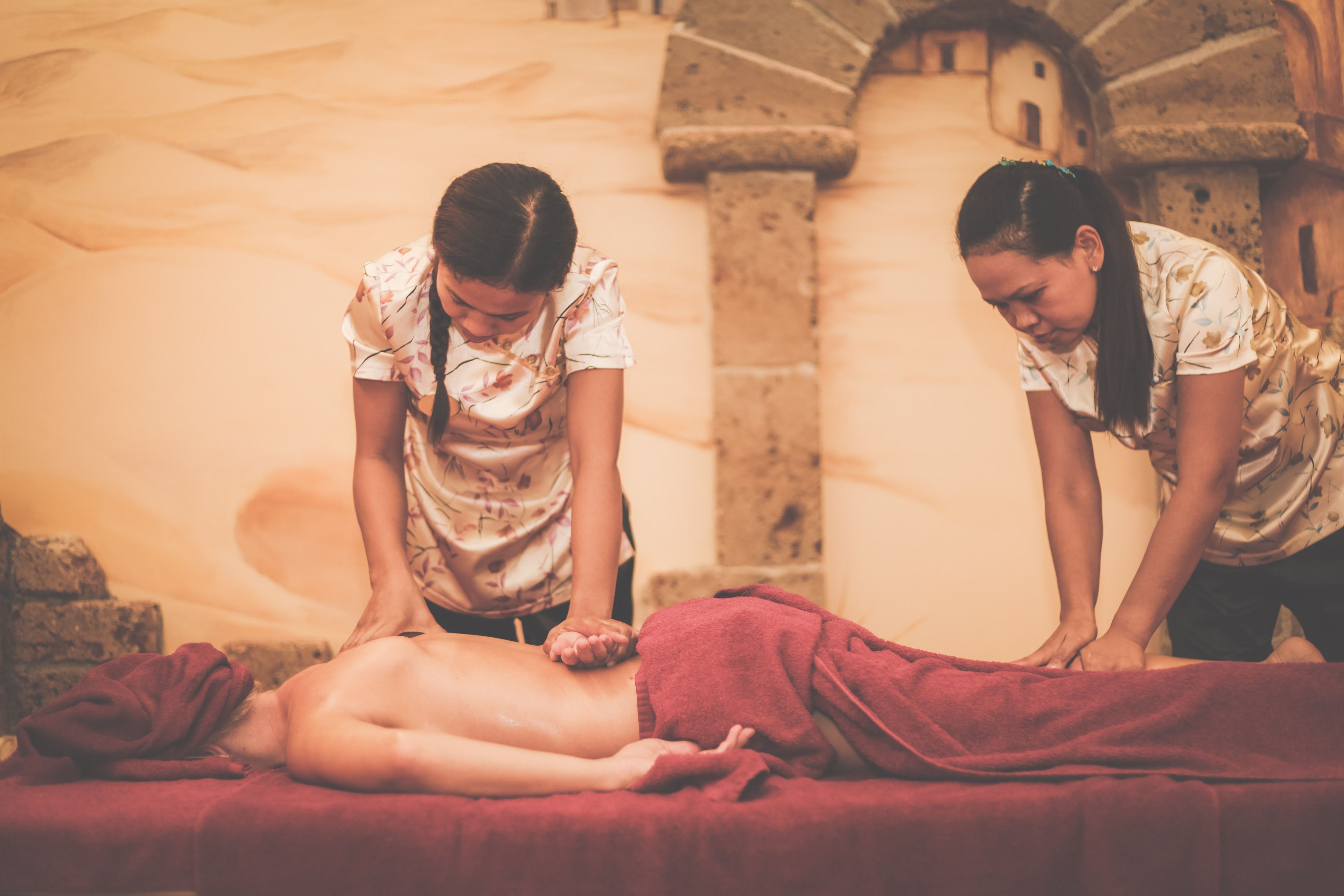 Eine Massageanwendung in einem Raum mit Steinwänden und einem roten Handtuch auf der Massageliege.