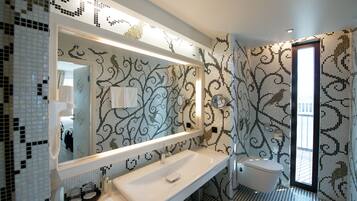 A modern bathroom with a large mirror, white sink, and toilet. The walls are decorated with a mosaic tile pattern.