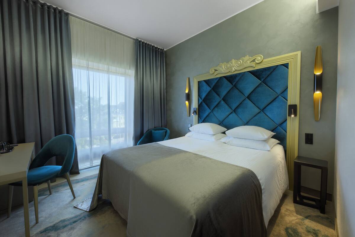 A hotel room with a large bed, a desk, a chair, and a view of the outdoors through the window.