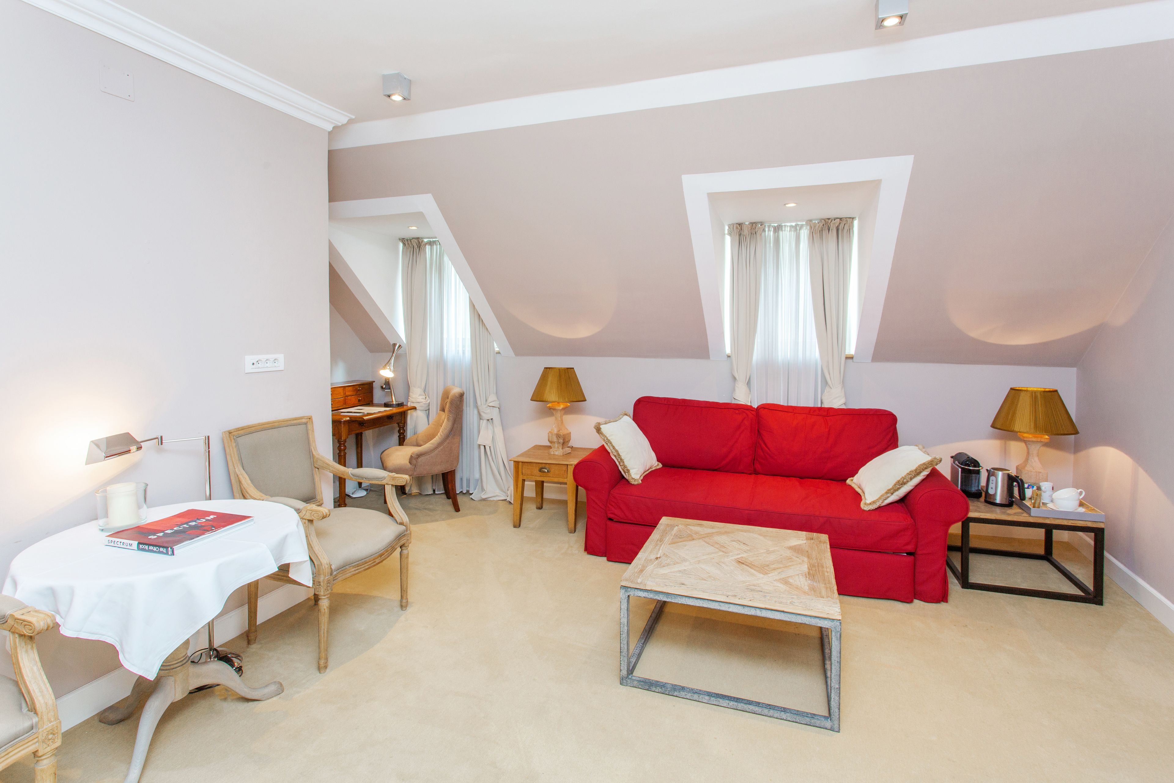 Ein gemütliches Wohnzimmer mit einem roten Sofa, einem kleinen Tisch und einem Sessel. Eine Lampe steht auf einem Beistelltisch und auf dem Tisch liegt ein Buch.