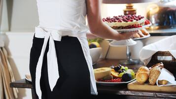 Iemand in een witte schort houdt een dessertbord vast met taart en fruit. Op de tafel staan broodjes en een mand.