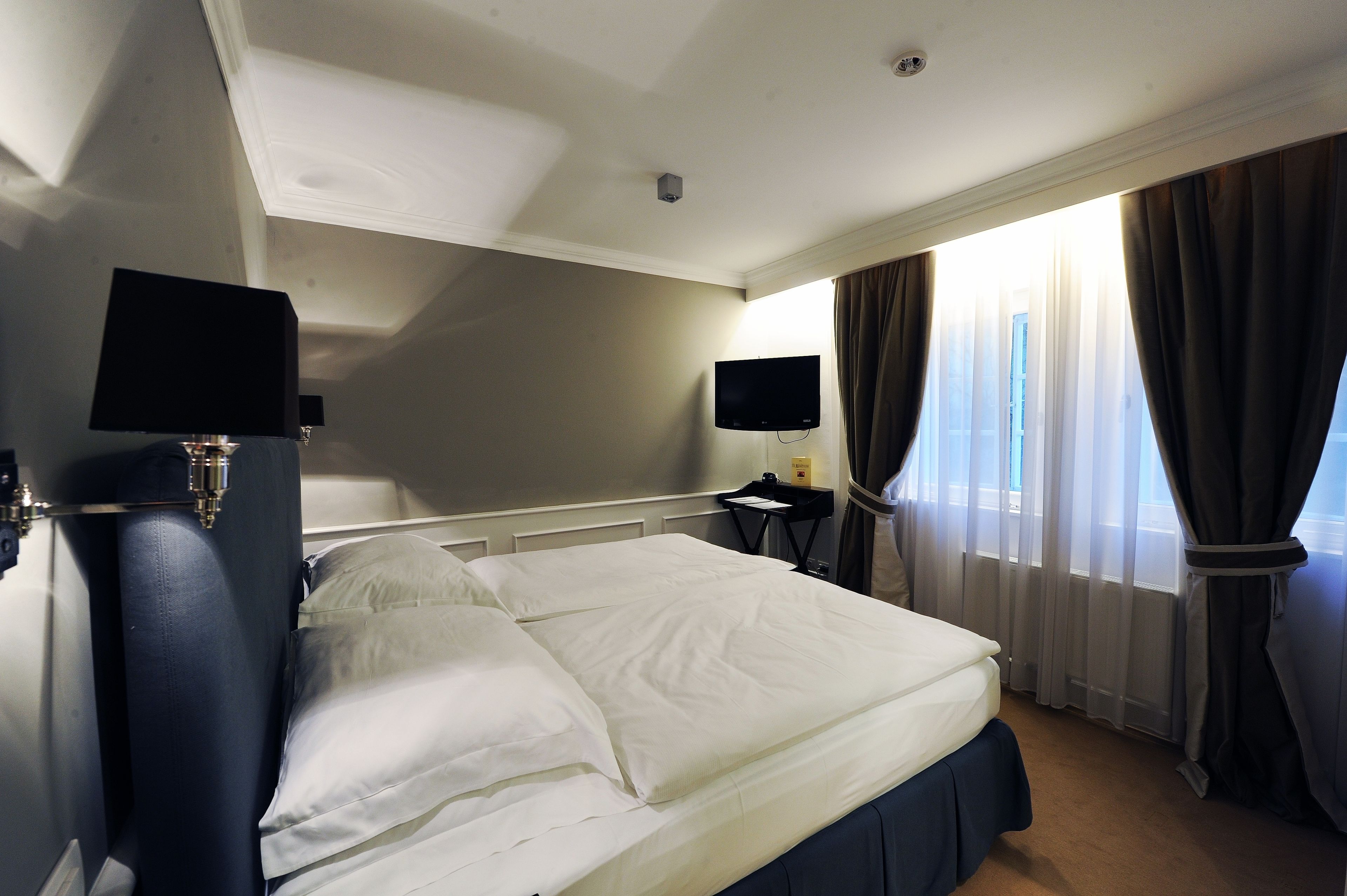 A hotel room with a bed, a flat-screen TV, a bedside table, and a window with curtains.