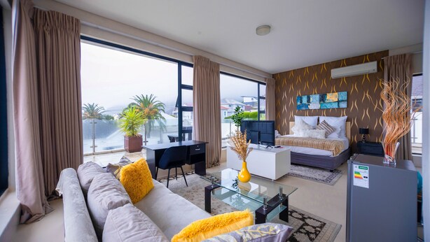 A modern living room with a sofa, coffee table, and a dining area. There is a large window with curtains, a potted plant, and a bed with a view. - Grande Kloof Boutique Hotel (Cape Town)
