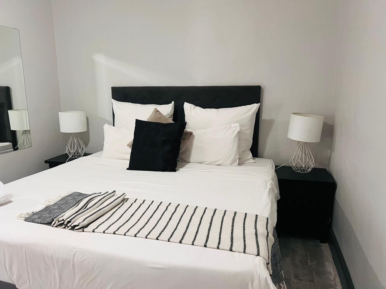 Ein ordentlich bezogenes Bett mit weißer Bettwäsche, schwarz-weißen Kissen und einer Streifen-Decke. Zwei Nachttische mit Lampen und ein Spiegel an der Wand sind vorhanden.