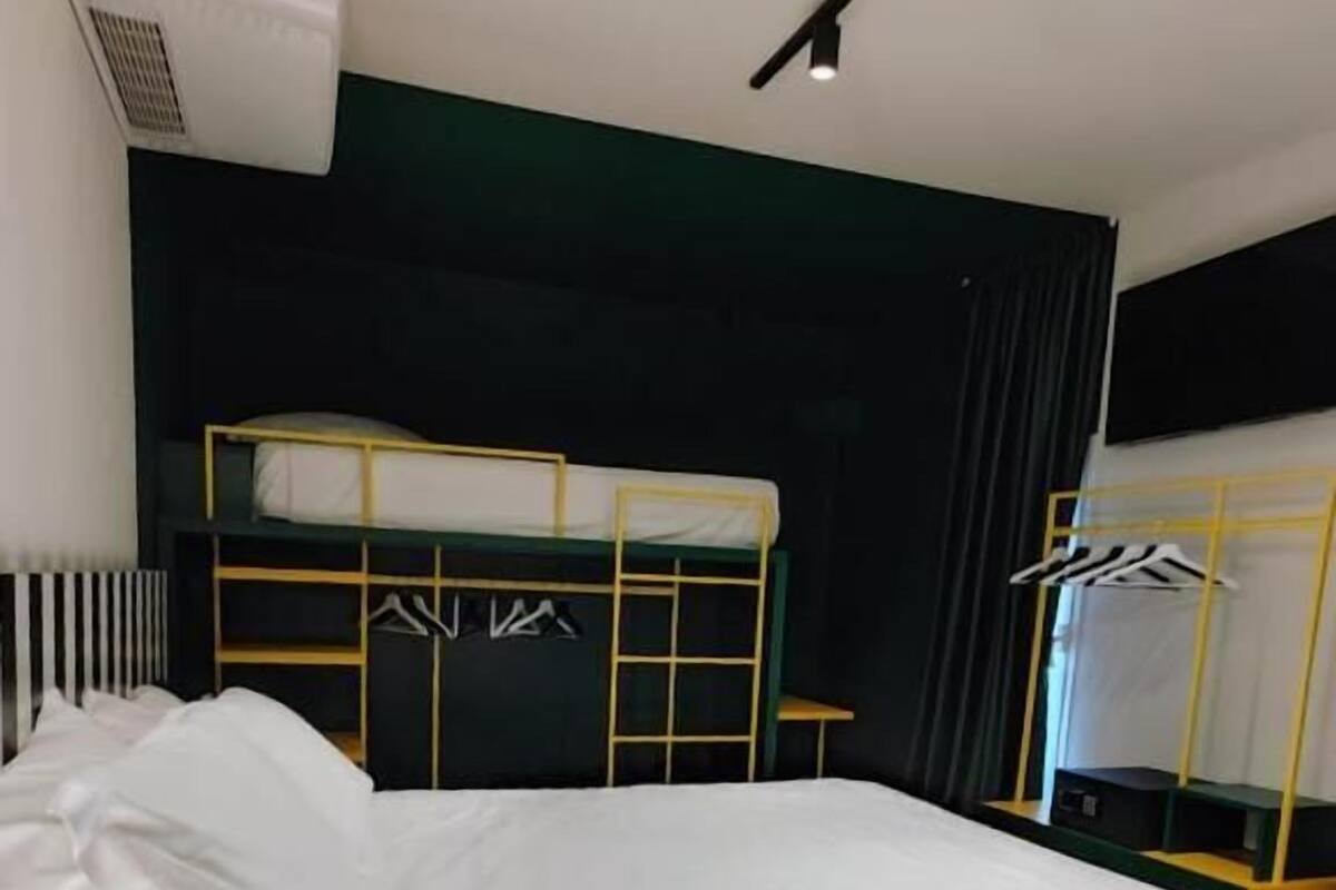 Un lit superposé avec un matelas blanc, une structure jaune et une tête de lit noire. Sur le mur, on aperçoit un appareil de climatisation blanc, ainsi qu'un rideau noir et une petite table avec une chaise.