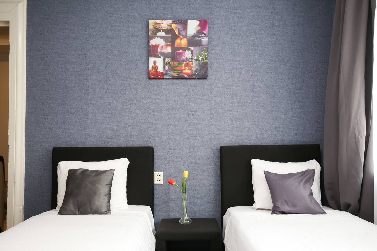 A hotel room with two beds, a small table with a vase of flowers, and a wall art piece above the beds.