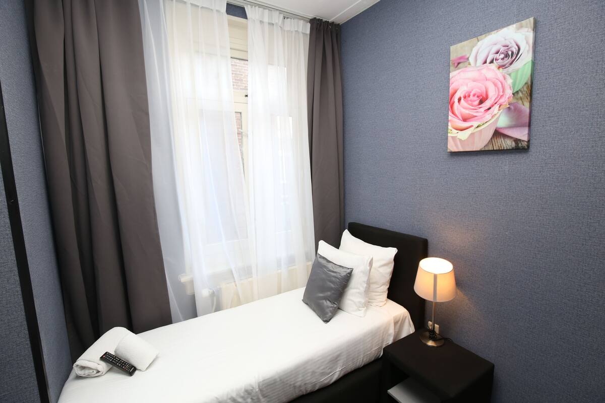 A hotel room with a bed, a nightstand with a lamp, a painting on the wall, and sheer curtains.