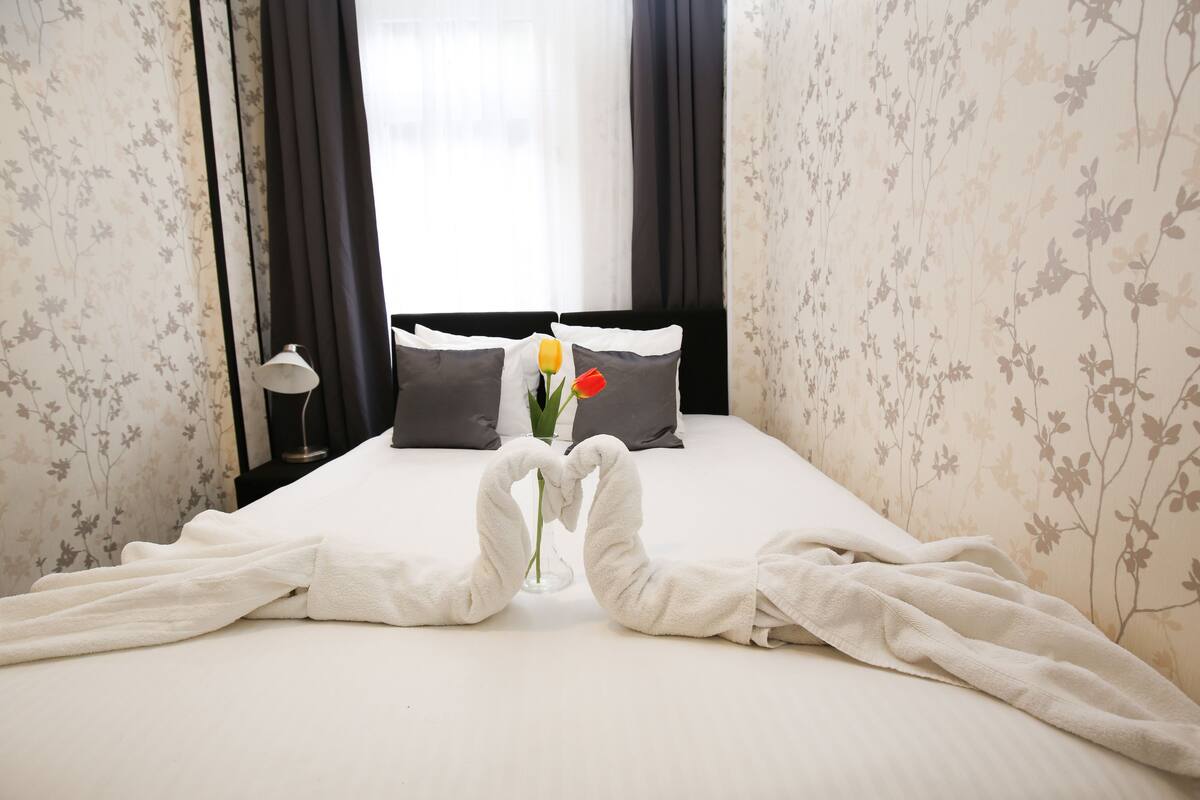 A neatly made bed with towels arranged in a heart shape, a vase with flowers, and a nightstand with a lamp.