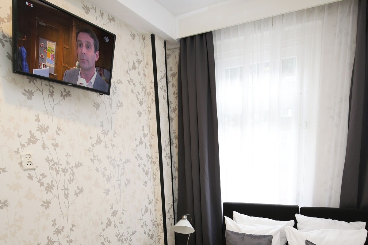 A hotel room with a bed, a TV mounted on the wall, a nightstand, and a window with curtains.