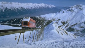 Un tren en una vía ferroviaria nevada en la montaña, con picos nevados al fondo.