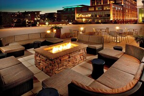 A rooftop lounge with a stone fire pit, cushioned seating, and a view of the cityscape.