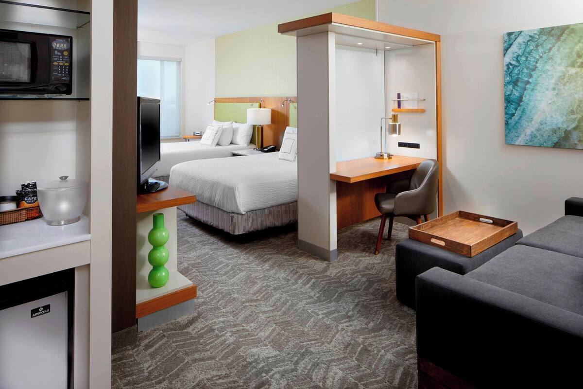 A hotel room with a bed, a desk, a chair, a microwave, a television, and a painting on the wall.