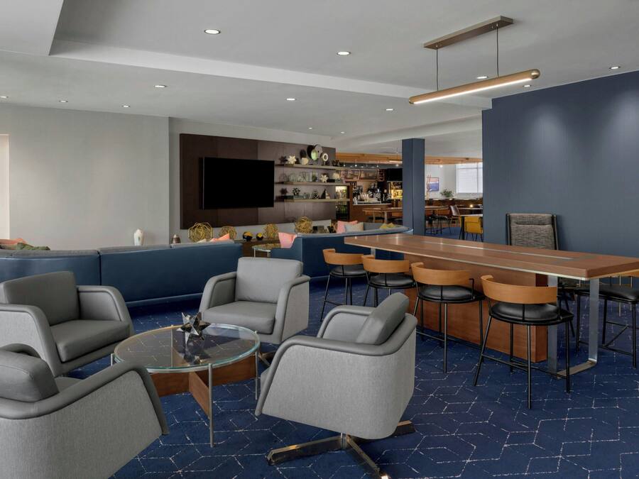 A modern hotel lobby with a blue carpet, a bar area with stools, and a seating area with sofas and chairs.