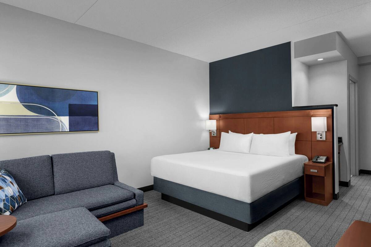 A hotel room with a bed, a gray sofa, a small table, and a painting on the wall.