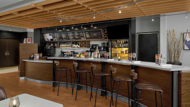 A modern bar area with a wooden ceiling, bar stools, a white countertop, and a menu board. - Courtyard Pittsburgh Airport Settlers Ridge (Pittsburgh)