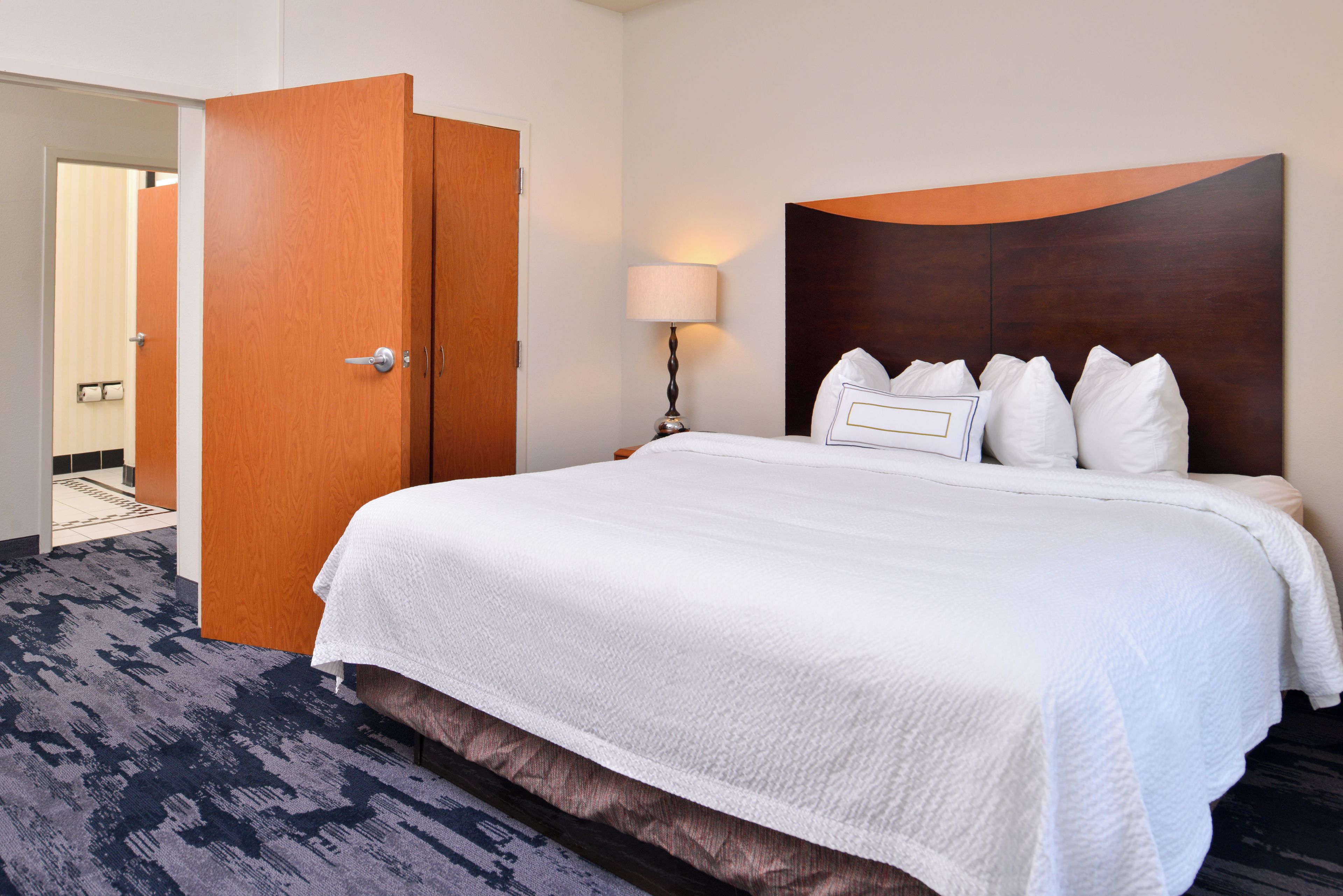 A hotel room with a large bed, a wooden door, a bedside lamp, and a carpeted floor.
