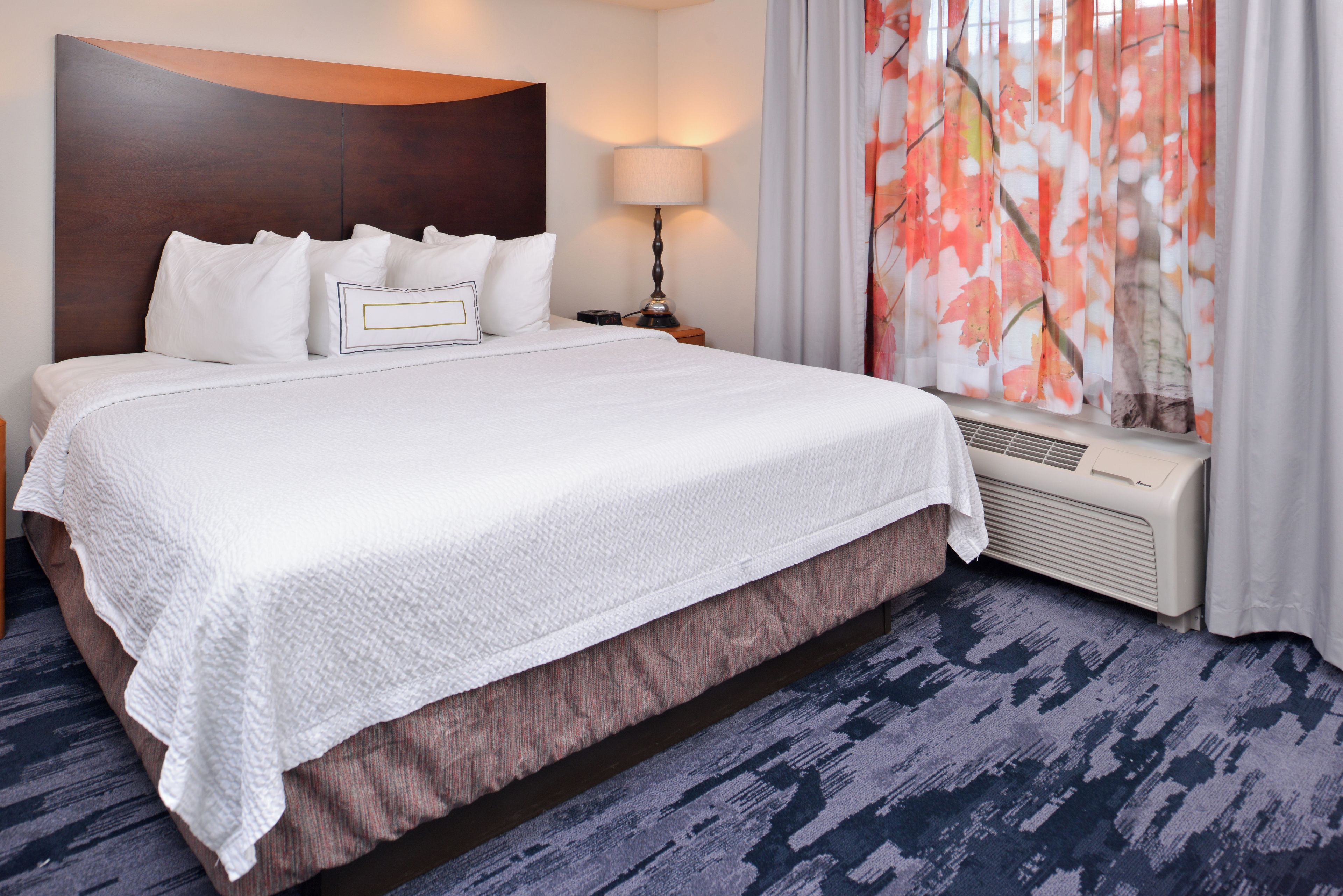 A hotel room with a large bed, a nightstand, a lamp, and a window with floral curtains.