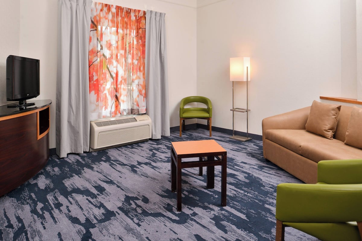 Habitación de hotel con televisor de pantalla plana, una mesita de madera, una silla verde, un sofá color beige y una lámpara de pie.