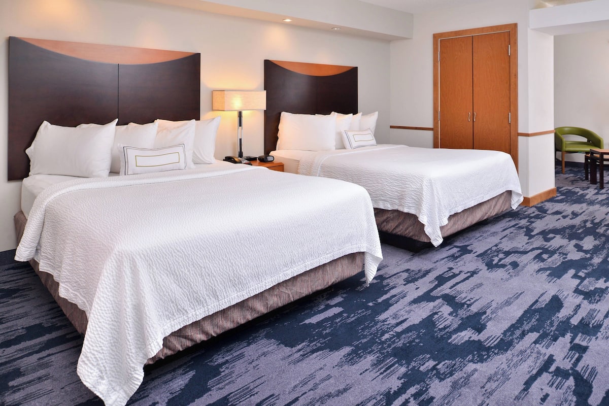Una habitación de hotel con dos camas, un armario de madera, una silla verde y una alfombra estampada.