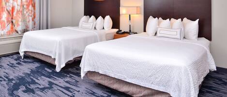 A hotel room with two beds, a nightstand, a lamp, and a view of a tree through a window.