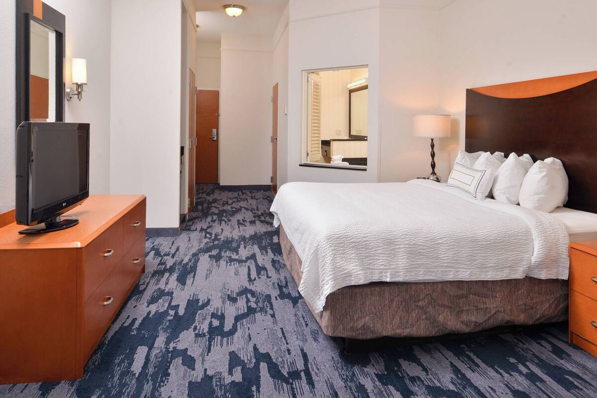 Una habitación de hotel con una cama, un televisor sobre un aparador de madera, un espejo y una lámpara.