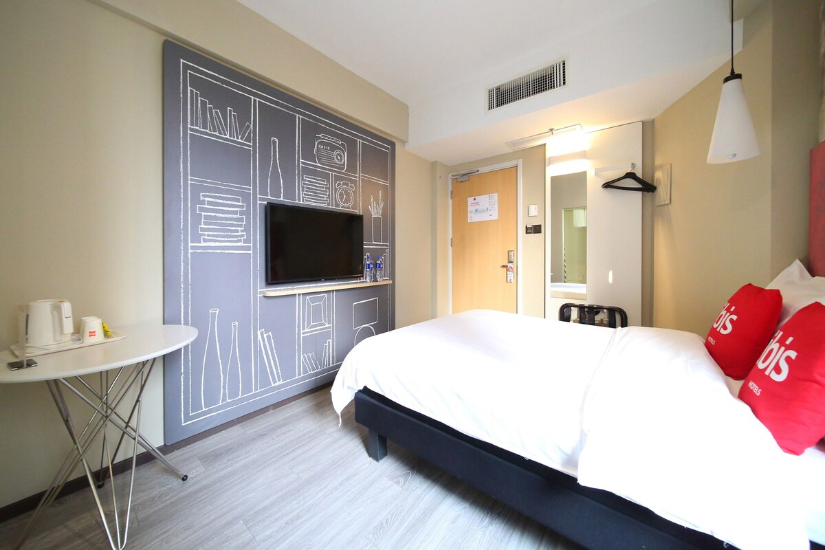 A hotel room with a bed, a TV mounted on the wall, a small table with a cup and a bottle, and a mirror.