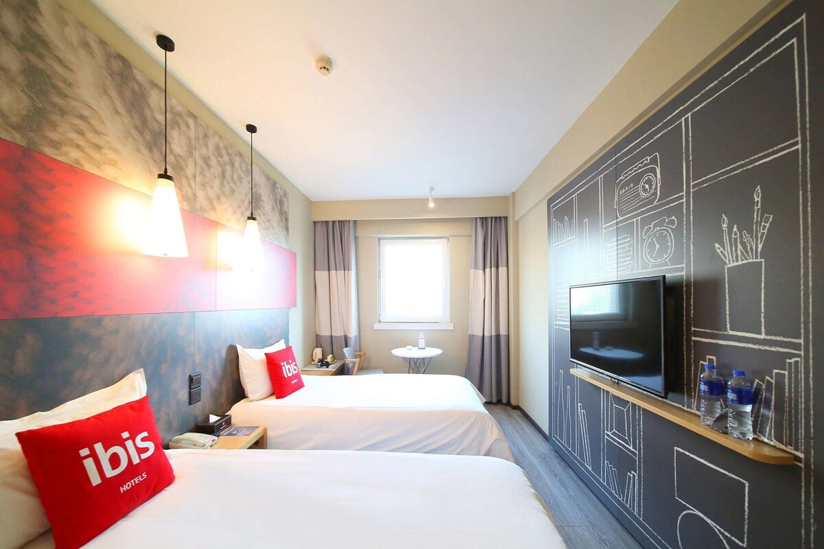 Une chambre d’hôtel avec deux lits, une télévision, une petite table et un mur recouvert d’un tableau noir où sont dessinés des motifs.