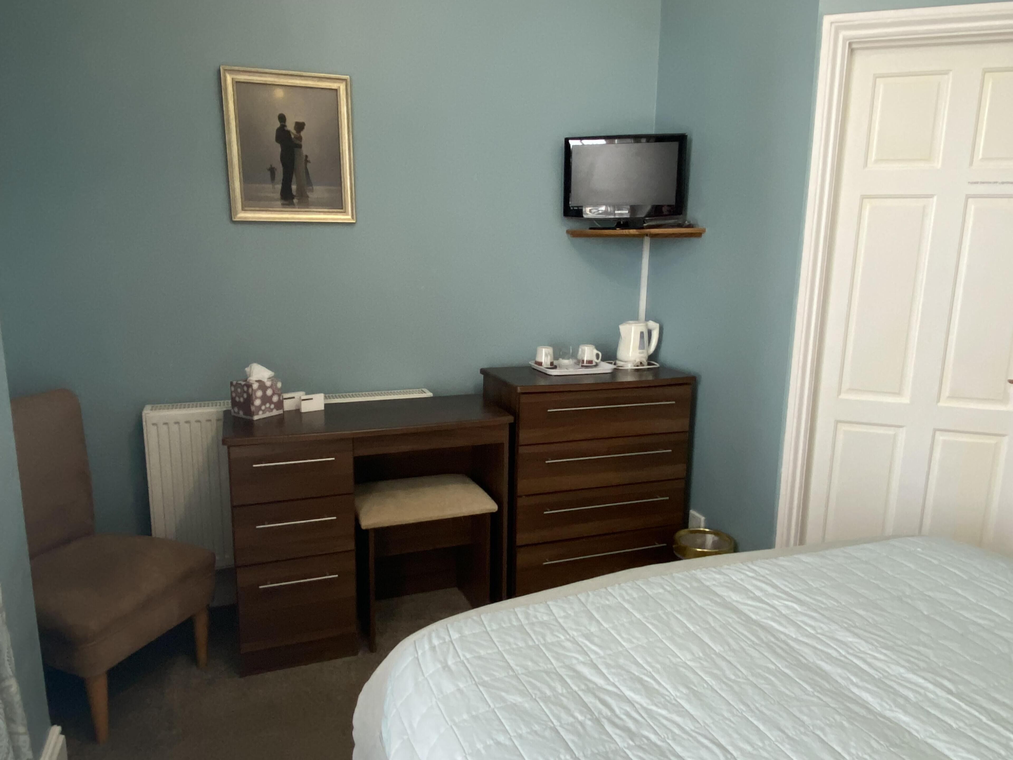Una camera da letto con un letto, una scrivania con un computer, una sedia, una TV su un supporto e un quadro appeso al muro.