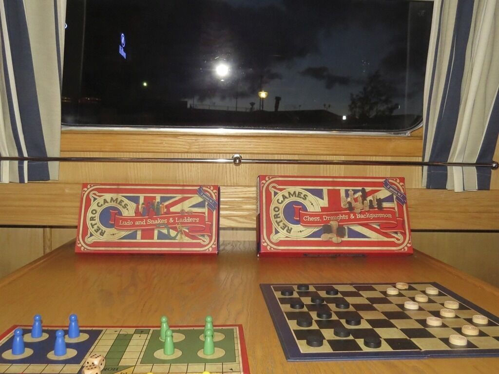 Dos juegos de mesa antiguos, 'Ludo y Serpientes y Escaleras' y 'Ajedrez, Damas y Nardos', sobre una mesa de madera con una ventana al fondo.