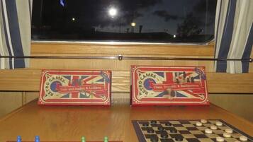 Dos juegos de mesa antiguos, 'Ludo y Serpientes y Escaleras' y 'Ajedrez, Damas y Nardos', sobre una mesa de madera con una ventana al fondo.