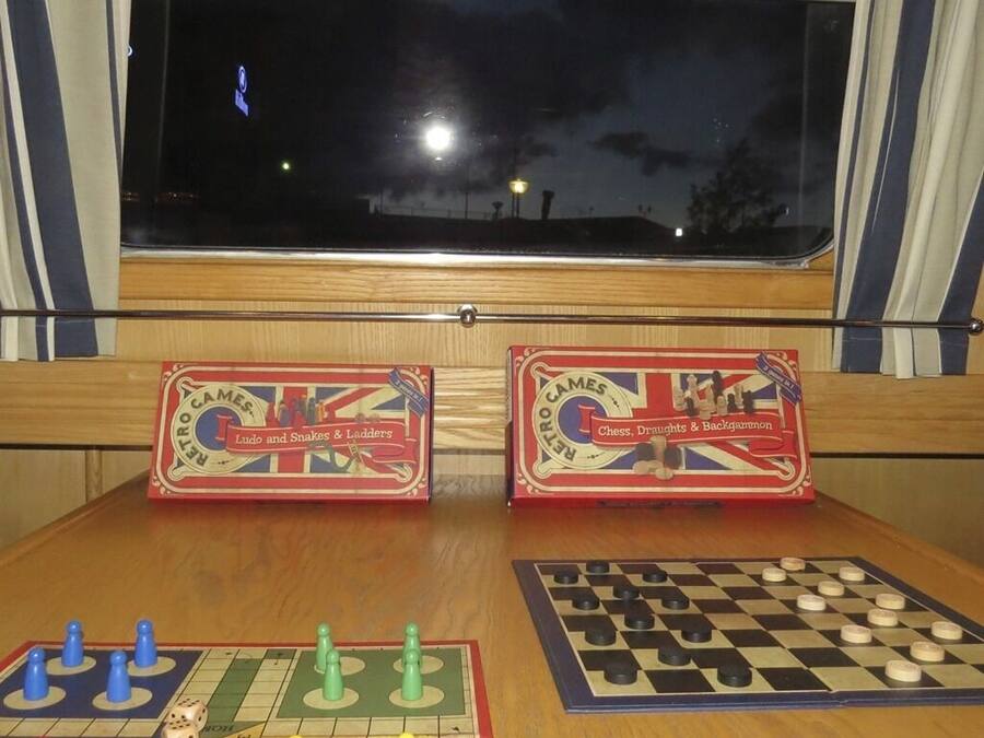 Dos juegos de mesa antiguos, 'Ludo y Serpientes y Escaleras' y 'Ajedrez, Damas y Nardos', sobre una mesa de madera con una ventana al fondo.