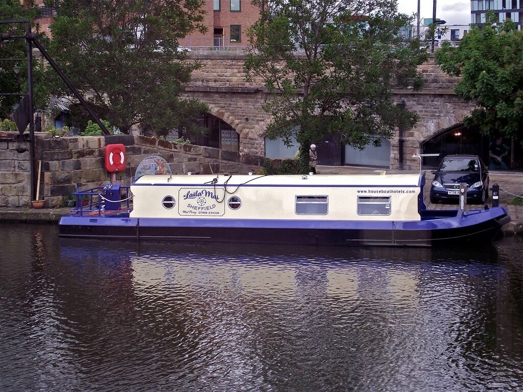 Een woonboot met een bord waarop 'Sheffield Houseboats' staat, afgemeerd bij een stenen brug.