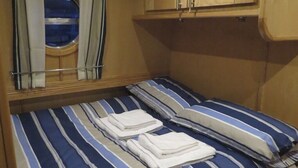Ein kleiner, kompakter Raum mit einem Einzelbett, gestreifter Bettwäsche und zwei Kissen. Es gibt ein Fenster mit Vorhängen und ein kleines rundes Fenster über dem Bett.