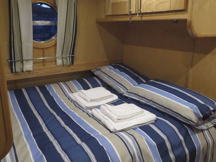 A small, compact room with a single bed featuring striped bedding and two pillows. There is a window with curtains and a small round window above the bed.
