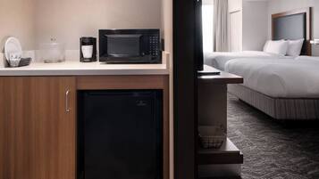 Une chambre d’hôtel dotée d’un coin cuisine comprenant un four à micro-ondes, une machine à café et un réfrigérateur. La chambre dispose d’un lit avec une literie blanche et une tête de lit, ainsi que d’un petit coin salon avec une chaise.
