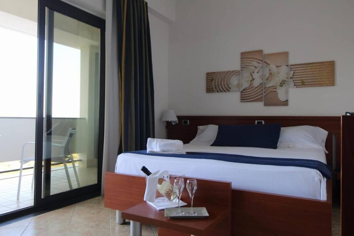 Ein Hotelzimmer mit Bett, Nachttisch mit Gläsern und einer Flasche, großem Fenster mit Vorhängen und Blick nach draußen.
