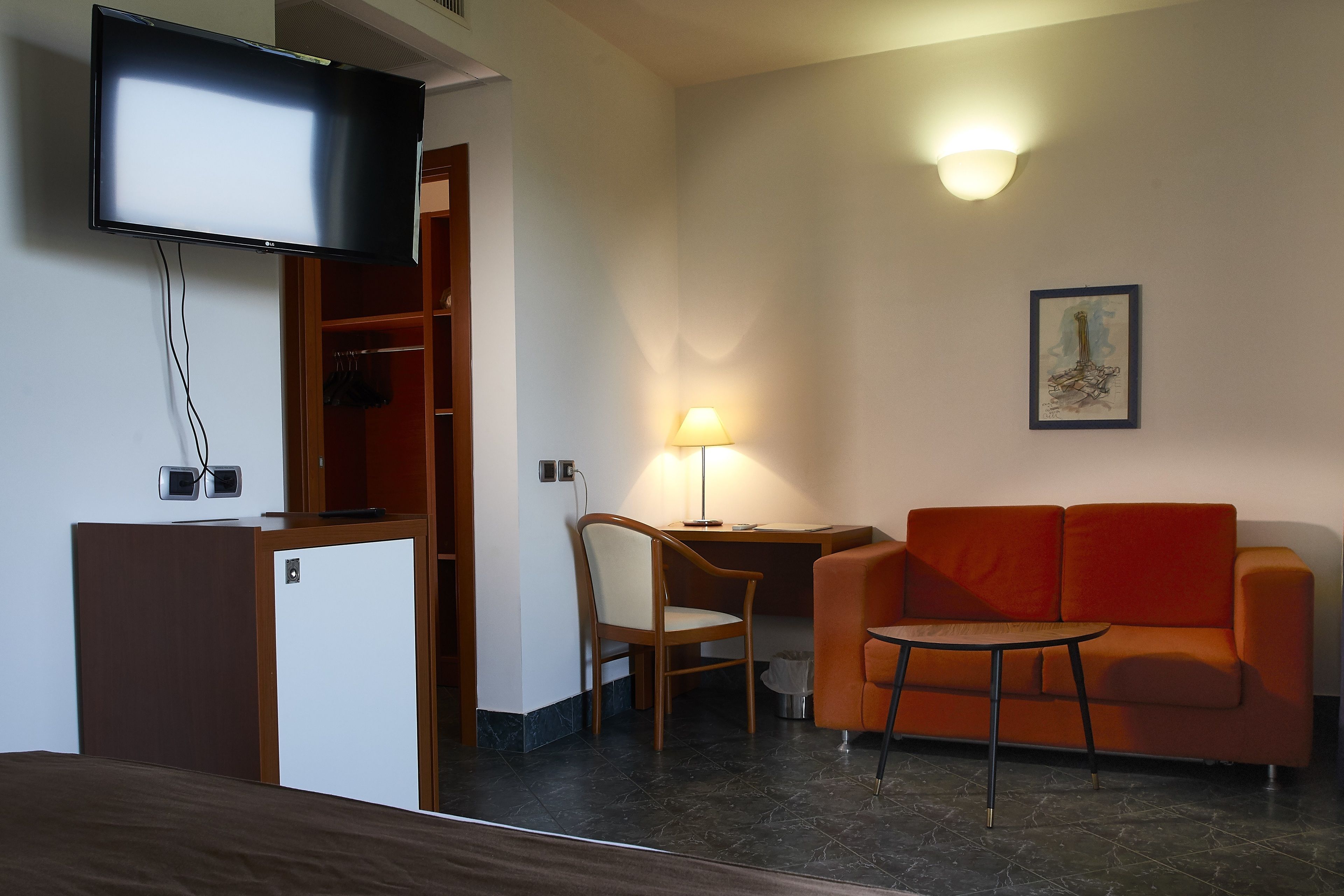 A hotel room with a bed, a TV mounted on the wall, a desk with a lamp, a sofa, and a chair.