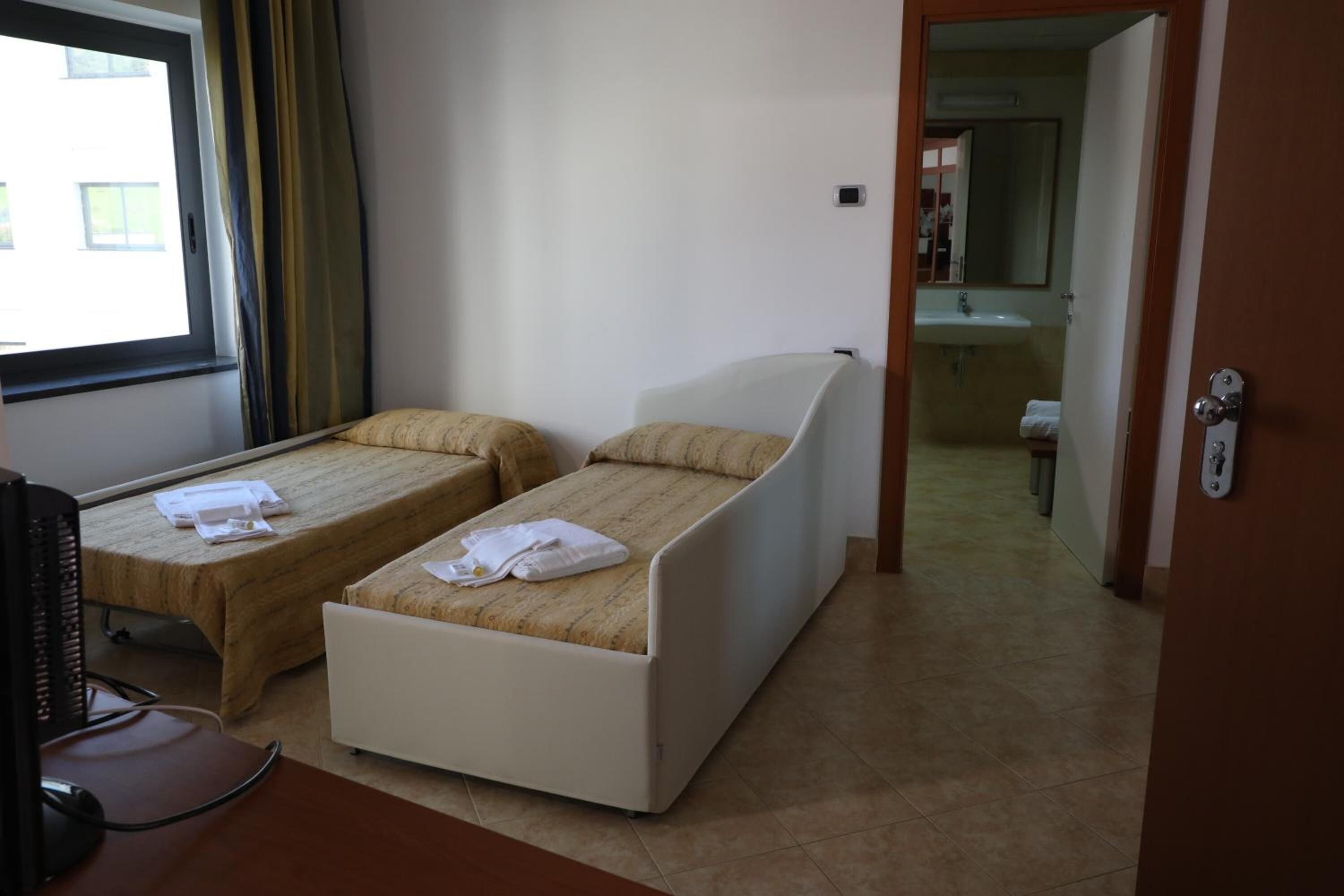 Habitación de hotel con dos camas individuales, un escritorio con una computadora, y un baño visible a través de una puerta abierta.