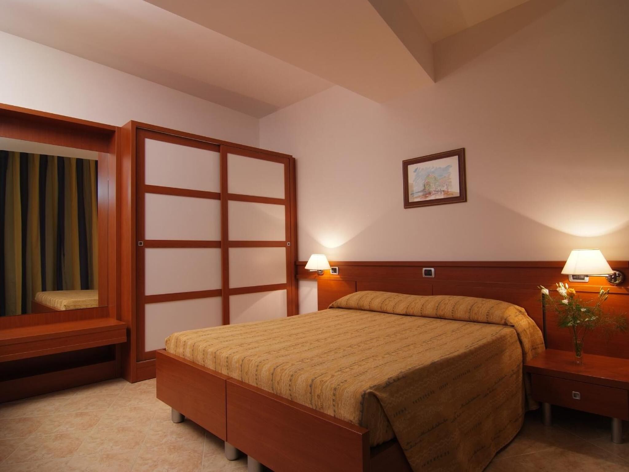 A hotel room with a bed, bedside lamps, a wooden headboard, a window with curtains, and a picture on the wall.