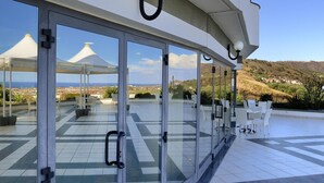 A modern outdoor patio with glass doors, white chairs, and a view of the sea and hills. - Park Hotel Mirabeau (Gasperina)