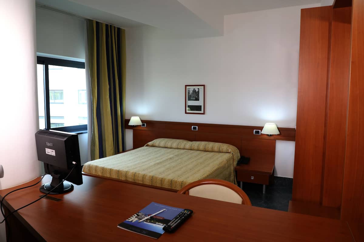 Ein Hotelzimmer mit Bett, Schreibtisch, Computer, Lampen und einem gerahmten Bild an der Wand.