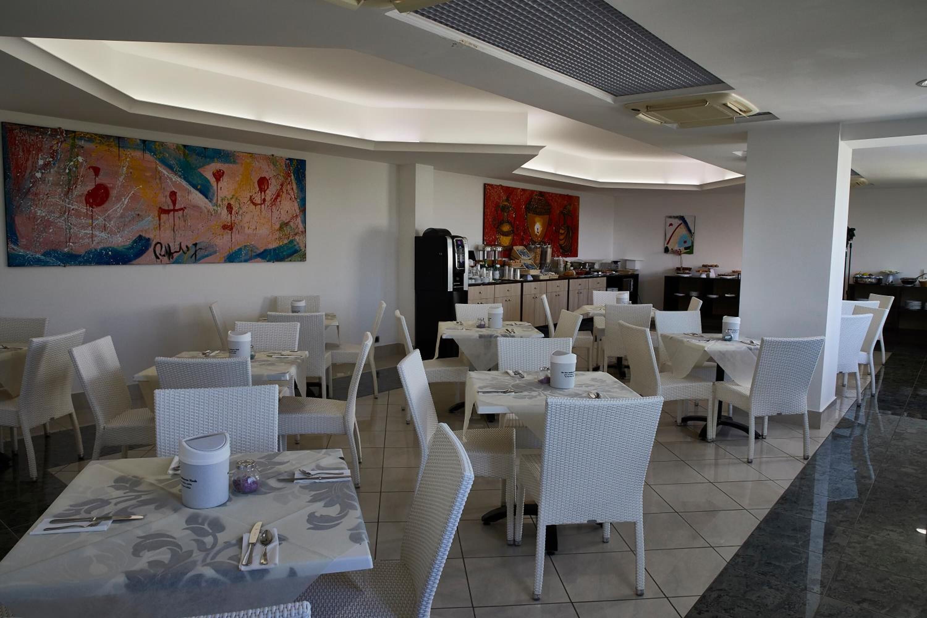 A restaurant with white wicker furniture, a large abstract painting on the wall, and a kitchen area in the background.