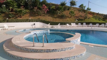 Ein Schwimmbecken mit Mosaikfliesenrand, umgeben von Liegestühlen und einem angelegten Garten mit verschiedenen Pflanzen und einer Hügelkulisse im Hintergrund.