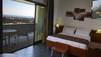A hotel room with a balcony, a bed with a wooden headboard, a bedside table with a lamp, and a wall-mounted artwork.
