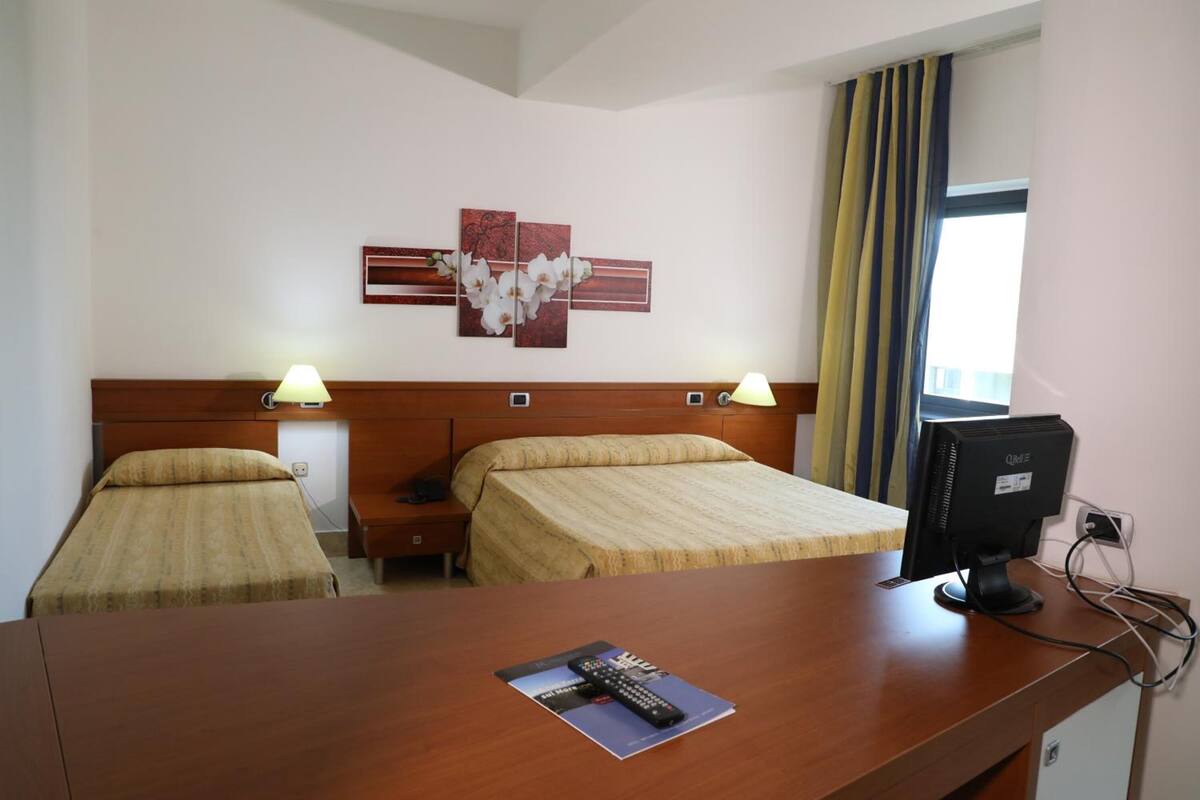 Ein Hotelzimmer mit zwei Betten, einem Schreibtisch mit Computer und einem Fenster mit Vorhängen.