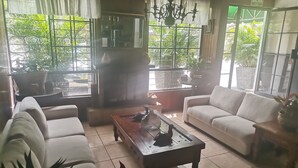 A spacious living room with a sofa, a coffee table, and a chandelier. Large windows with curtains allow natural light to fill the room. - Hotel Alicante San Salvador (Antiguo Cuscatlán)