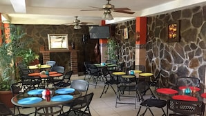 An outdoor patio area with multiple tables and chairs, stone walls, and a ceiling fan. - Hotel Alicante San Salvador (Antiguo Cuscatlán)